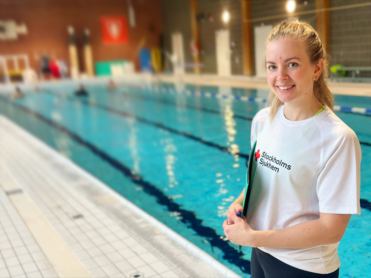 En medarbetare från Stockholms Sjukhem står vid bassängkanten i en simhall och ler mot kameran. I bakgrunden syns personer som tränar i vattnet.”