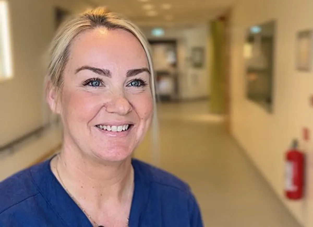 Leende kvinna klädd i blått i sjukhuskorridor.