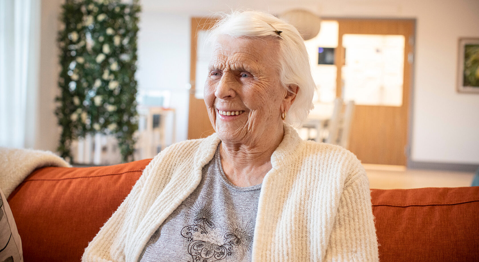 Leende äldre kvinna med vitt hår sitter på en orange soffa i ett ljust vardagsrum.