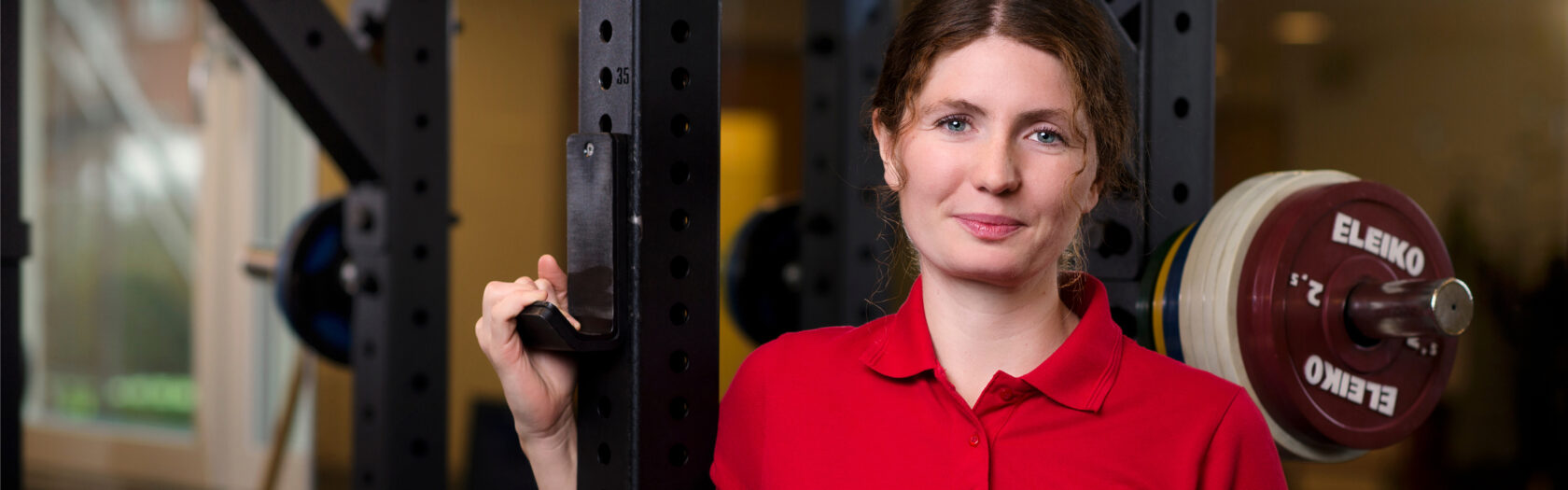 Kvinna i röd pikétröja står i gym bredvid skivstång med viktskivor och tittar mot kameran.