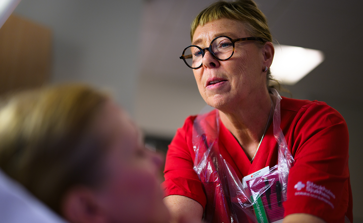 Sjuksköterska i röd uniform pratar med patient i sjukhussäng, fokus på vårdpersonalens ansikte.