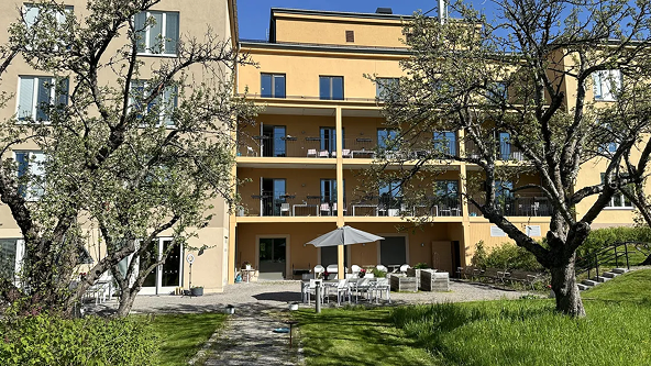 Gult flerbostadshus med balkonger, trädgård och utemöbler under ett parasoll. Två träd i förgrunden.