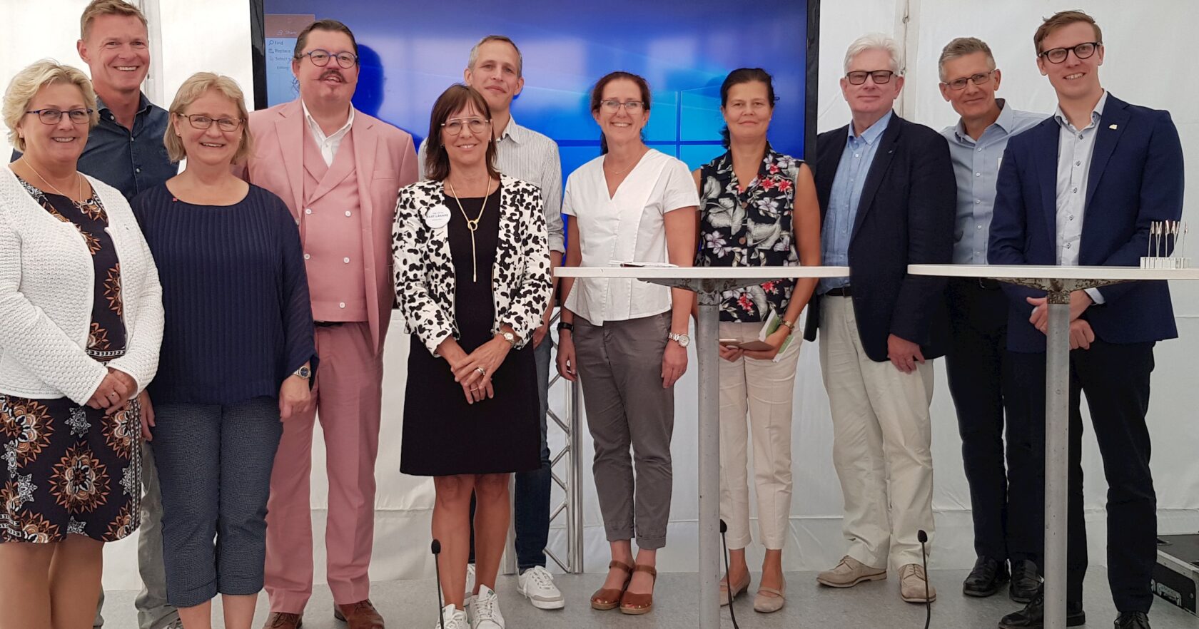 En grupp på elva personer står inomhus framför en stor skärm och ler mot kameran.