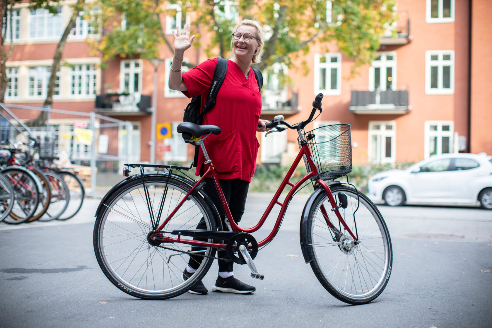 Leende person i röd tröja står bredvid en röd cykel på en stadsgata och vinkar mot kameran.