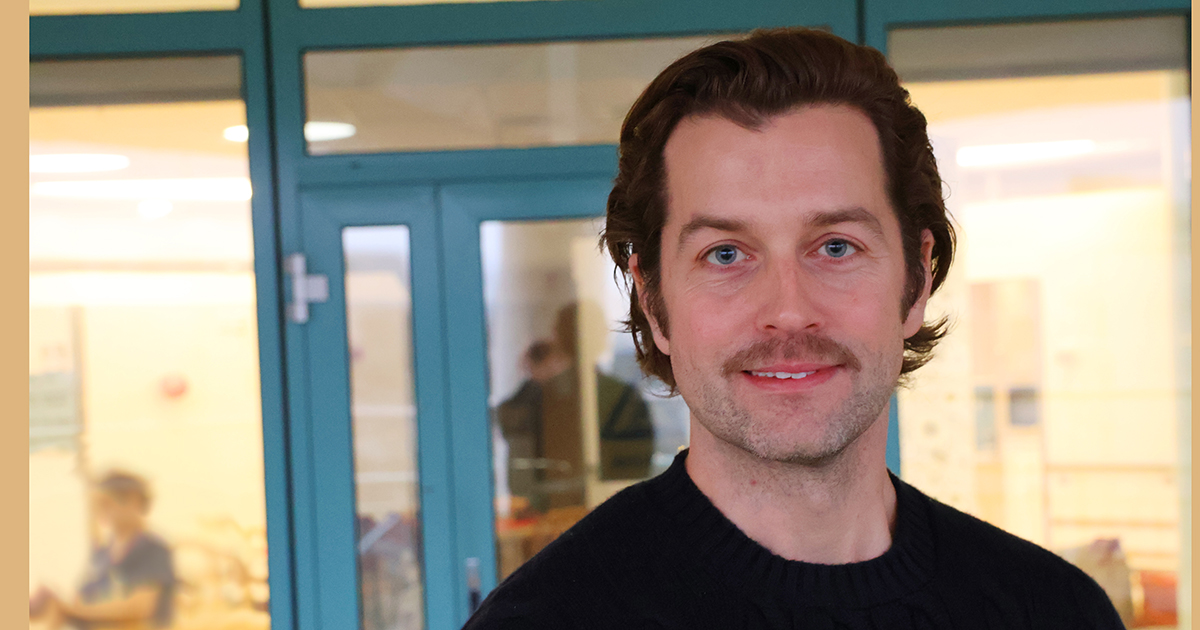 Man med brunt hår och mustasch ler framför glasdörrar inomhus.