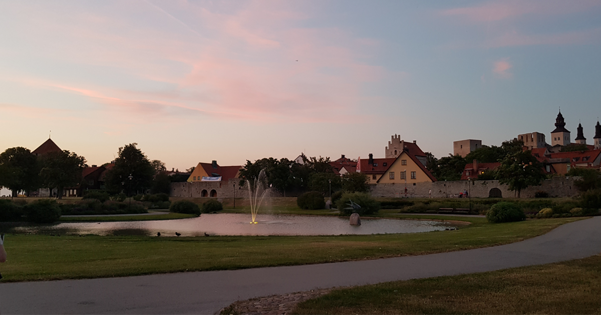 Park med damm och fontän i Visby, omgiven av gamla stadsmuren och byggnader i solnedgång.