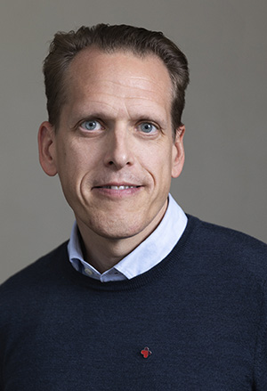 Man med kort brunt hår, ljus hy och blå tröja med skjorta, ler mot kameran mot neutral bakgrund.