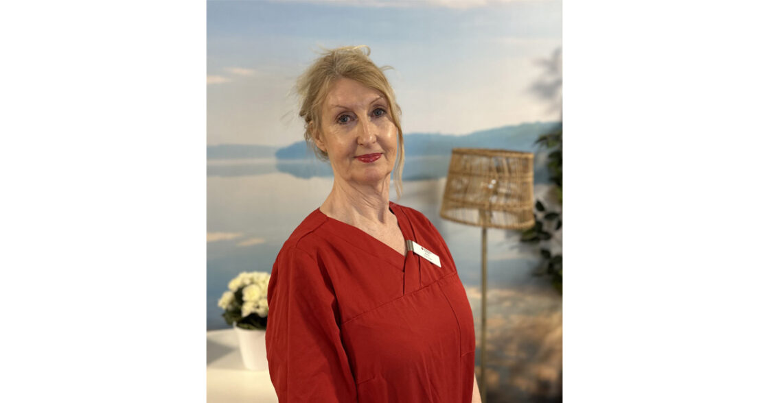 Kvinna i röd arbetsklädsel står inomhus med blommor och lampa i bakgrunden.