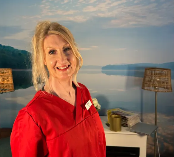 Leende kvinna i röd arbetsklädsel står framför en väggmålning med naturmotiv, inomhusmiljö med lampor.