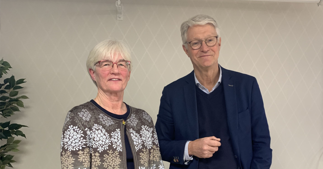 Två äldre personer med grått hår och glasögon står bredvid varandra inomhus mot ljus vägg.