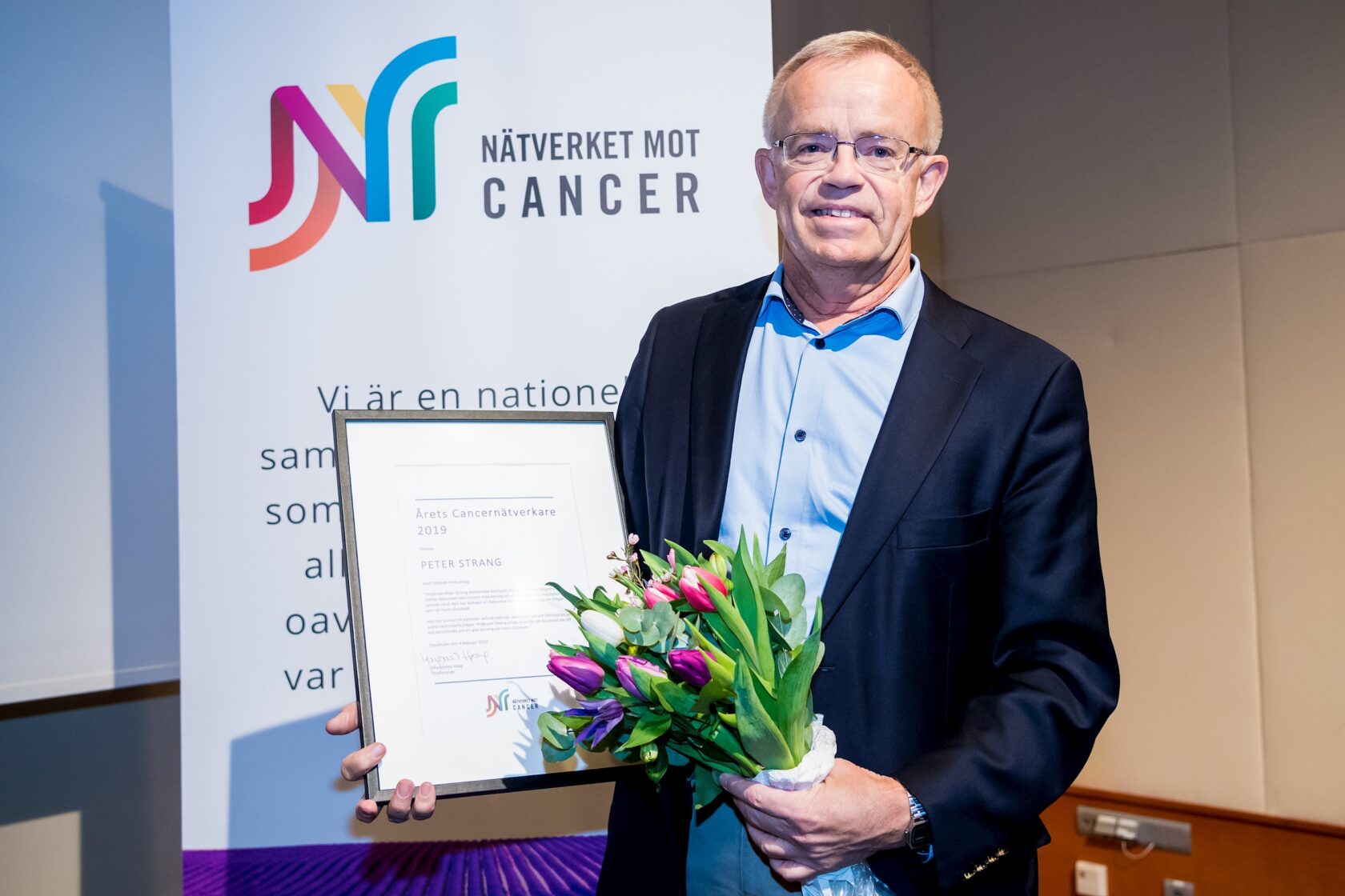 Man i kostym håller diplom och blombukett framför skylt med texten "Nätverket mot cancer".