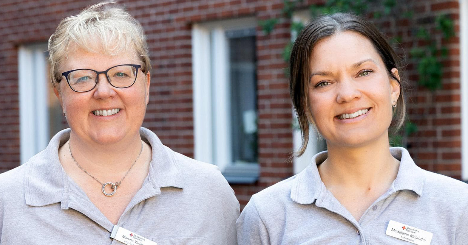 Två kvinnor i ljusgråa pikétröjor står utomhus framför en tegelvägg och ler mot kameran.