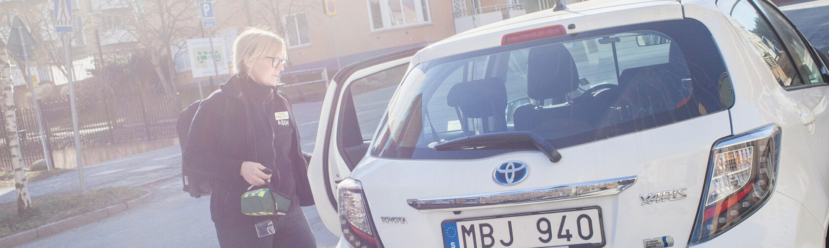 Kvinna i arbetskläder står vid en vit Toyota Yaris med öppen dörr på en gata i bostadsområde.