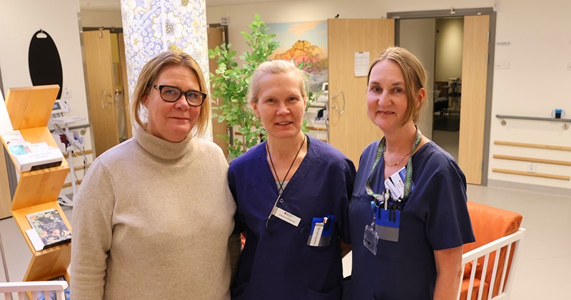Tre kvinnor, två i blå vårdkläder och en i beige tröja, står tillsammans på ett sjukhus eller vårdcentral.