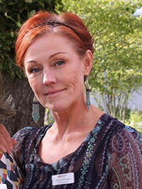 Kvinna med rött hår och stora örhängen står utomhus, bär mönstrad blus och namnbricka.