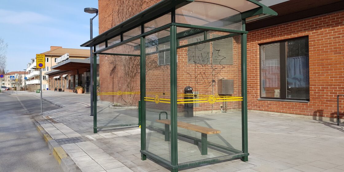 Busshållplats med glasväggar och bänk, bredvid en byggnad med röd tegelfasad. Soligt väder.