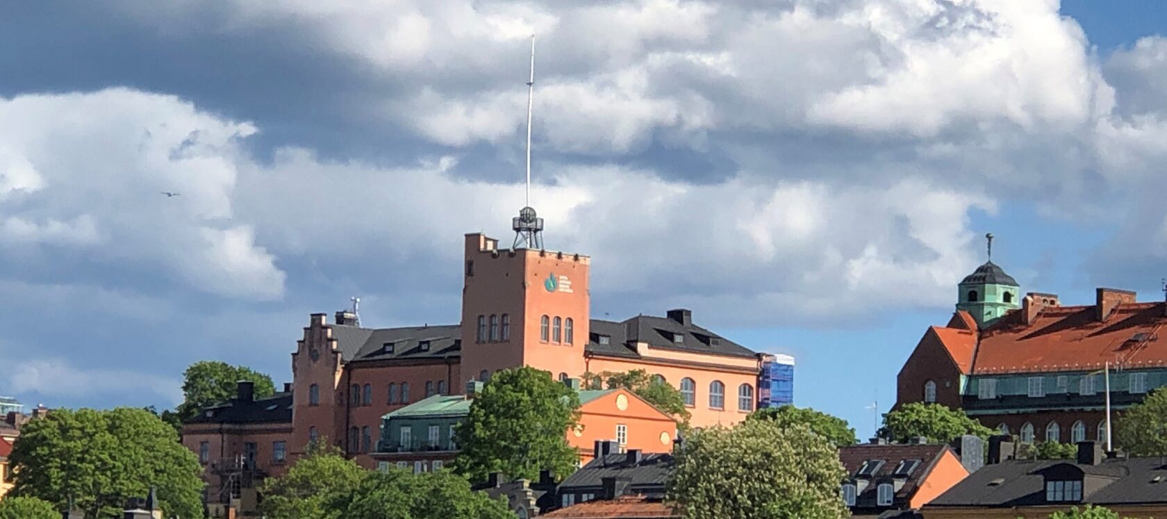 Röd tegelbyggnad med torn, gröna träd, hustak och blå himmel med moln i bakgrunden.