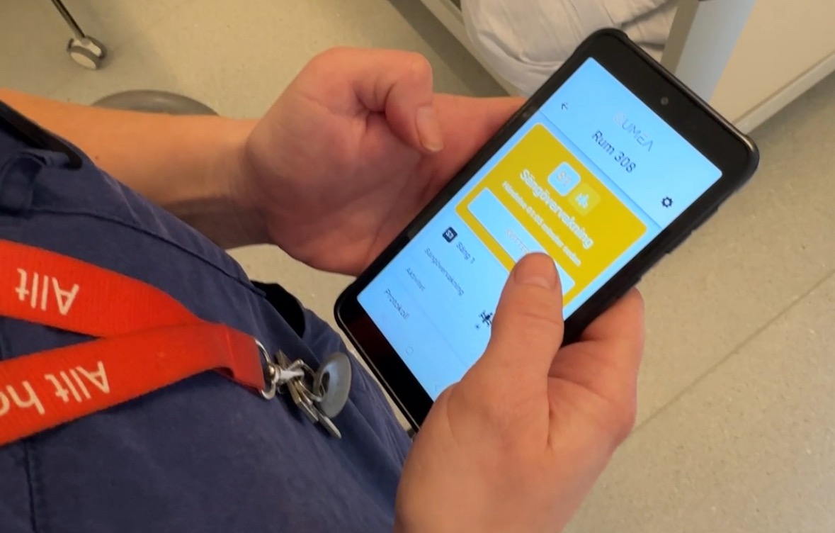Person håller en mobiltelefon med en app öppen, bär röd nyckelrem med texten "Allt".