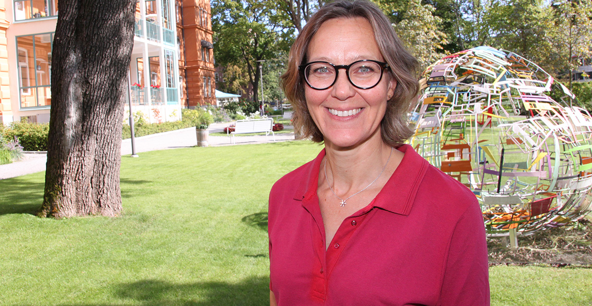 Leende kvinna med glasögon står utomhus på gräsmatta, konstverk och träd syns i bakgrunden.