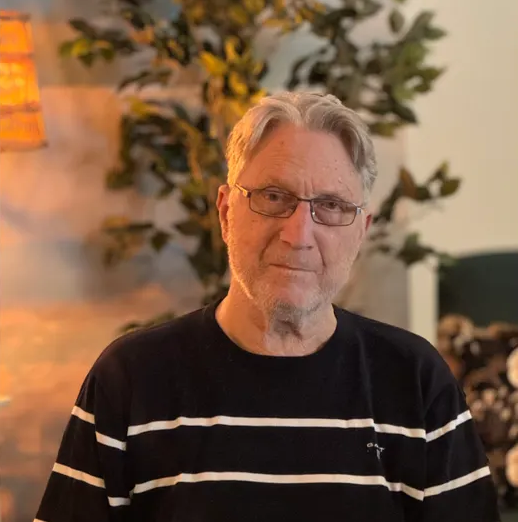 Äldre man med glasögon och randig tröja sitter inomhus, växt och lampa i bakgrunden.