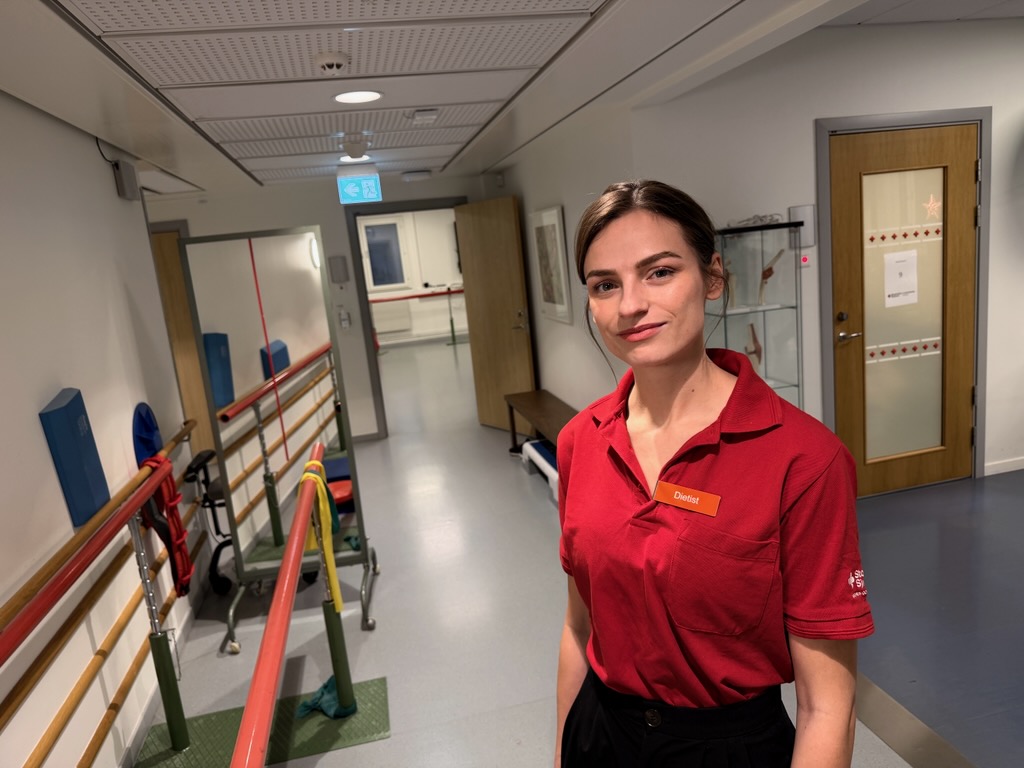 Kvinna i röd skjorta med namnskylt "Dietist" står i vårdkorridor med gånghjälpmedel.