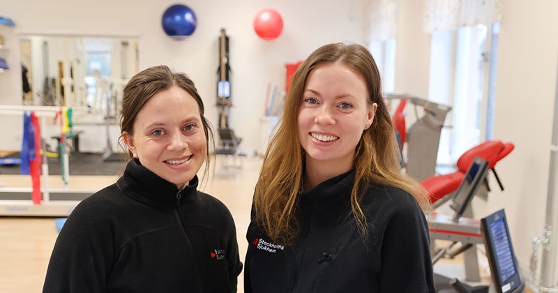 Två kvinnor i träningskläder står och ler i ett ljust gym med träningsutrustning i bakgrunden.