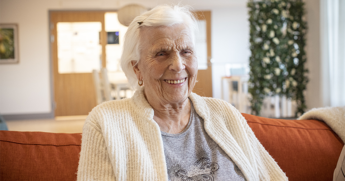 En äldre kvinna med vitt hår sitter leende på en orange soffa i ett ljust rum.