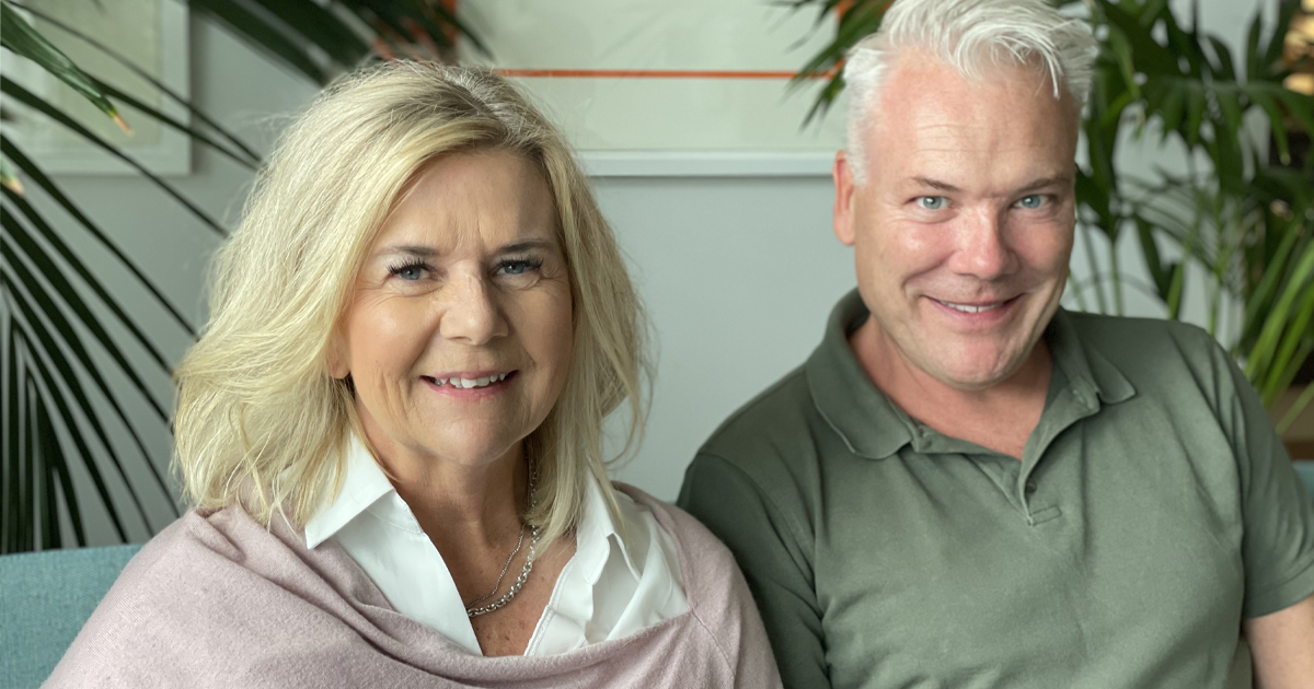 Två leende vuxna personer sitter bredvid varandra inomhus med gröna växter i bakgrunden.