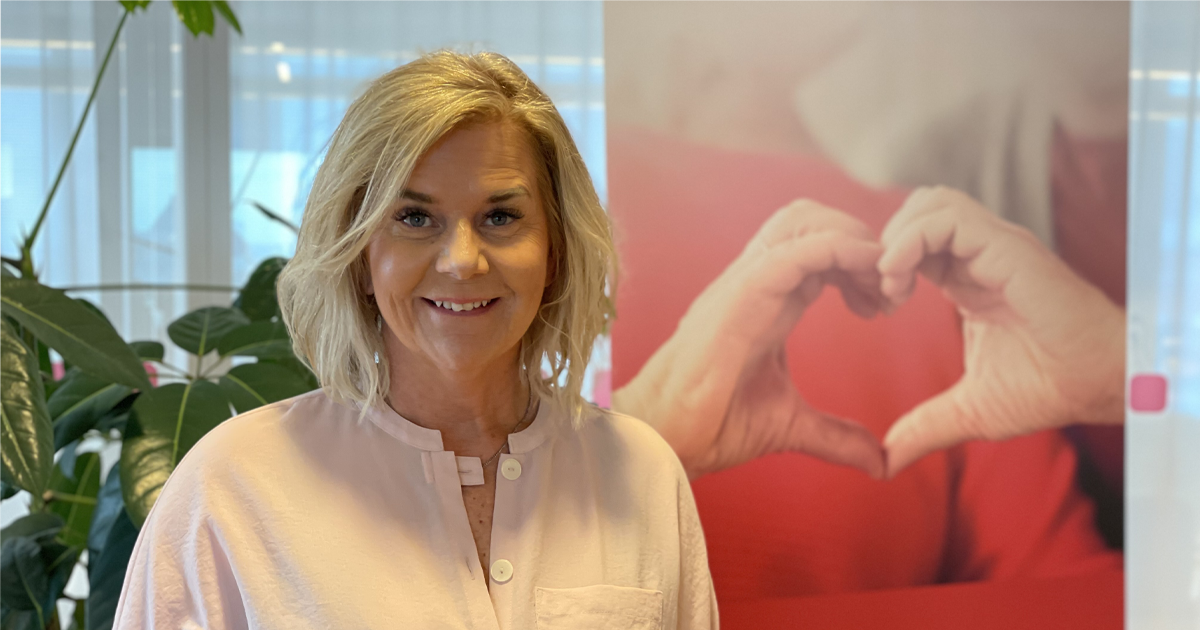 Leende kvinna i ljusrosa skjorta står inomhus. Bakom henne syns händer som formar ett hjärta.