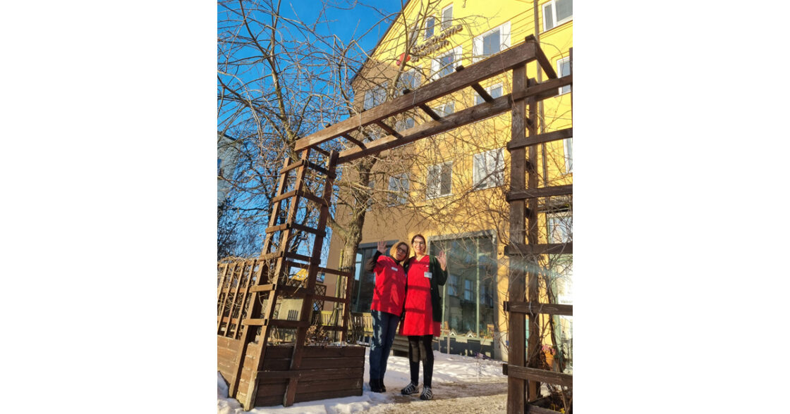 Två personer i röda jackor vinkar under en träpergola framför en gul byggnad, vintermiljö.