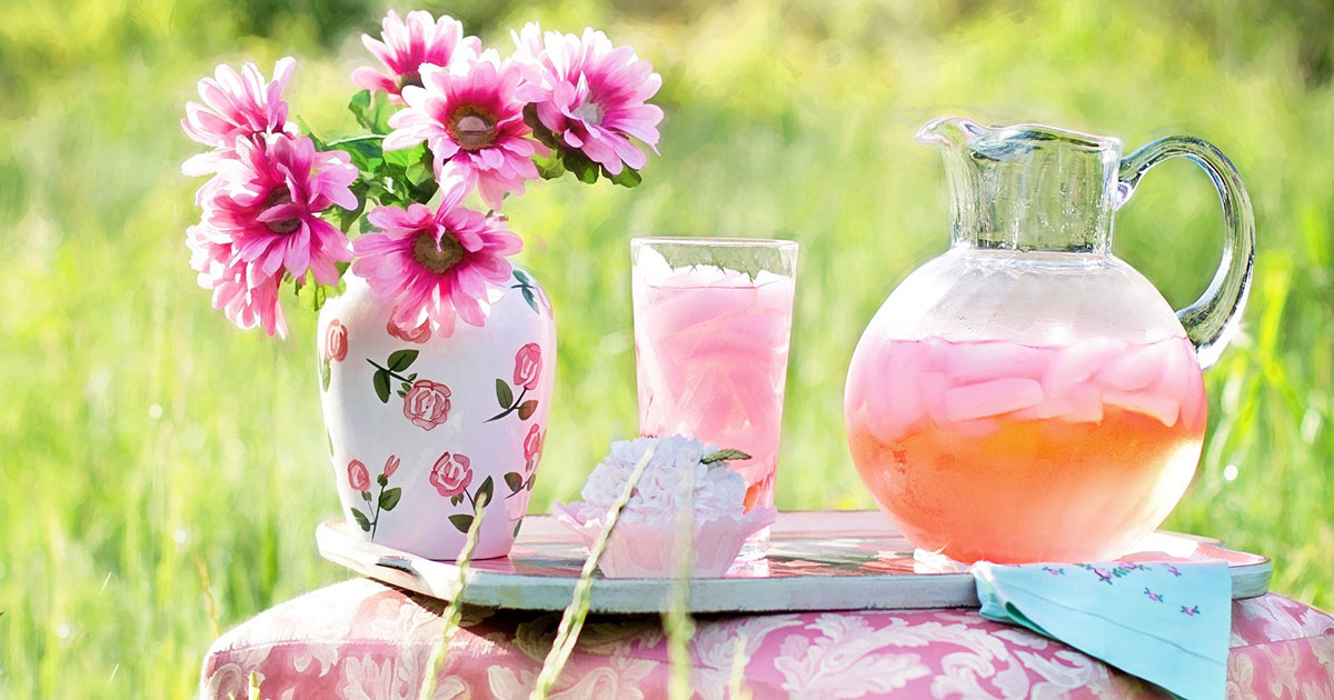Vas med rosa blommor, kanna och glas med rosa dryck och is, cupcake och servett på ett bord utomhus.