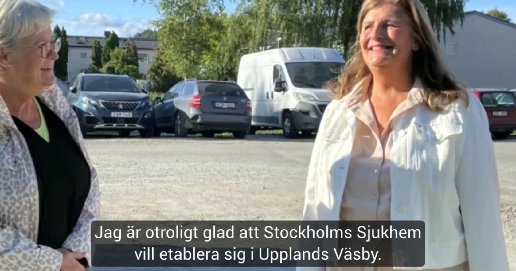 Två kvinnor utomhus, leende mot varandra, med bilar och träd i bakgrunden. Undertext på svenska visas.