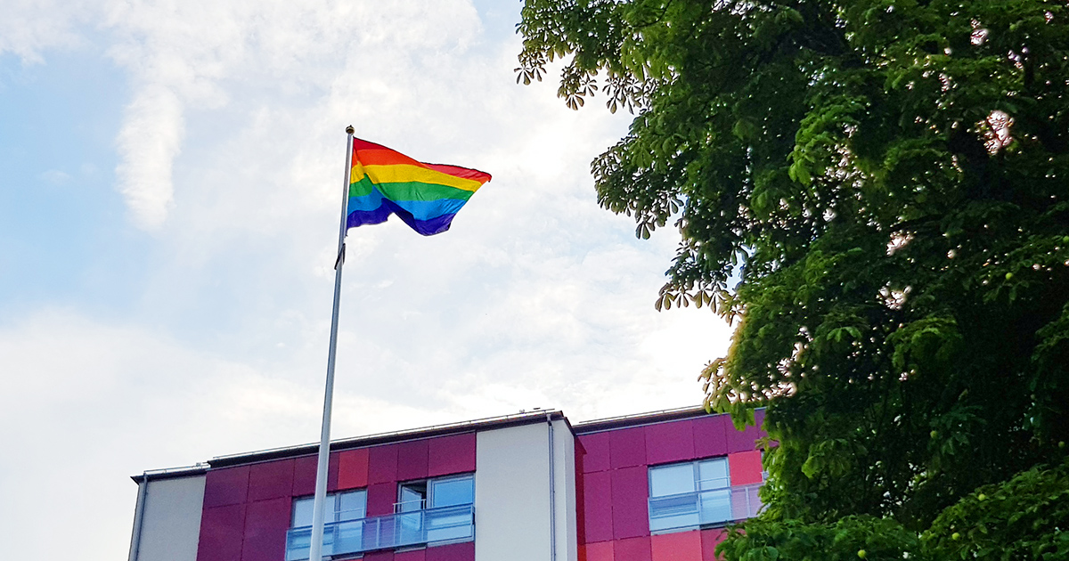 En regnbågsflagga vajar på en flaggstång framför en modern byggnad och ett träd.