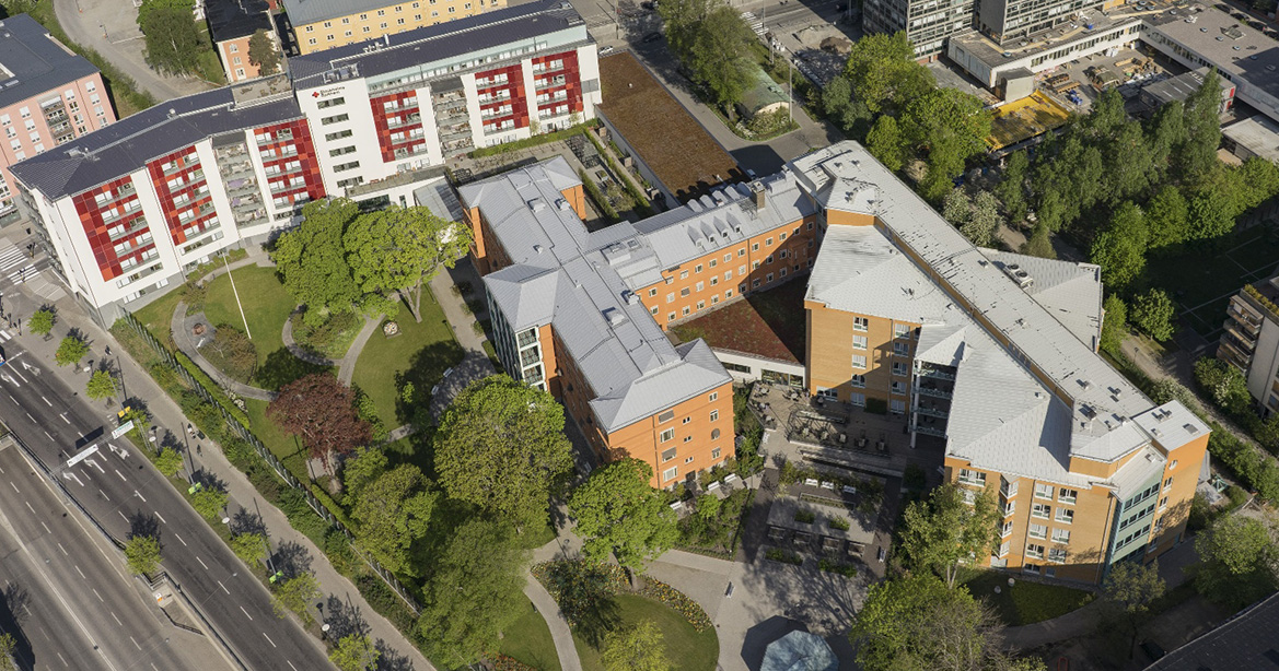 Fågelvy över bostadshus och grönområden i stadsmiljö med vägar och träd runt byggnaderna.