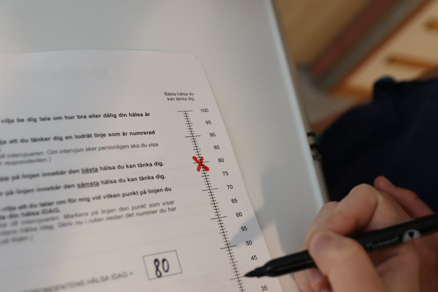 En hand håller en penna och fyller i en hälsoskala på papper, markerar siffran 80 med ett rött kryss.