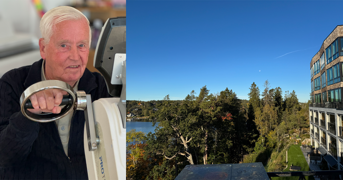 Äldre man tränar på träningsmaskin inomhus, bredvid utsikt över skog och sjö från en byggnad i solsken.