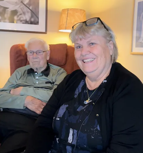 En äldre man och kvinna sitter tillsammans i ett varmt upplyst rum och ler mot kameran.