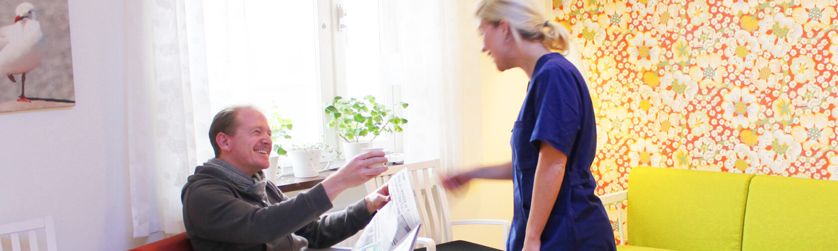 En man sitter leende med en tidning och får en kopp från en kvinna i vårdkläder i ett ljust rum.