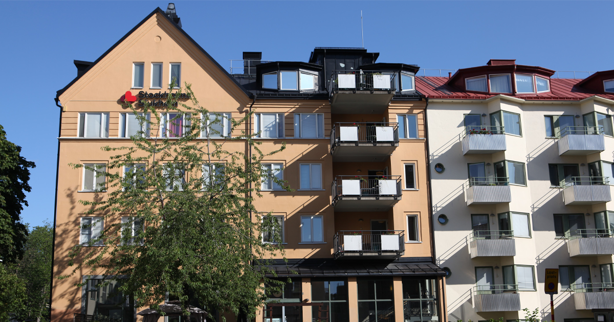 Två flerfamiljshus med balkonger i solsken, träd framför, blå himmel i bakgrunden.