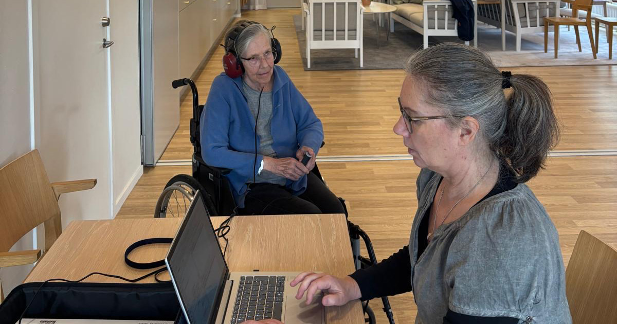 Två kvinnor vid ett bord, en sitter i rullstol med hörlurar, den andra arbetar vid en laptop.
