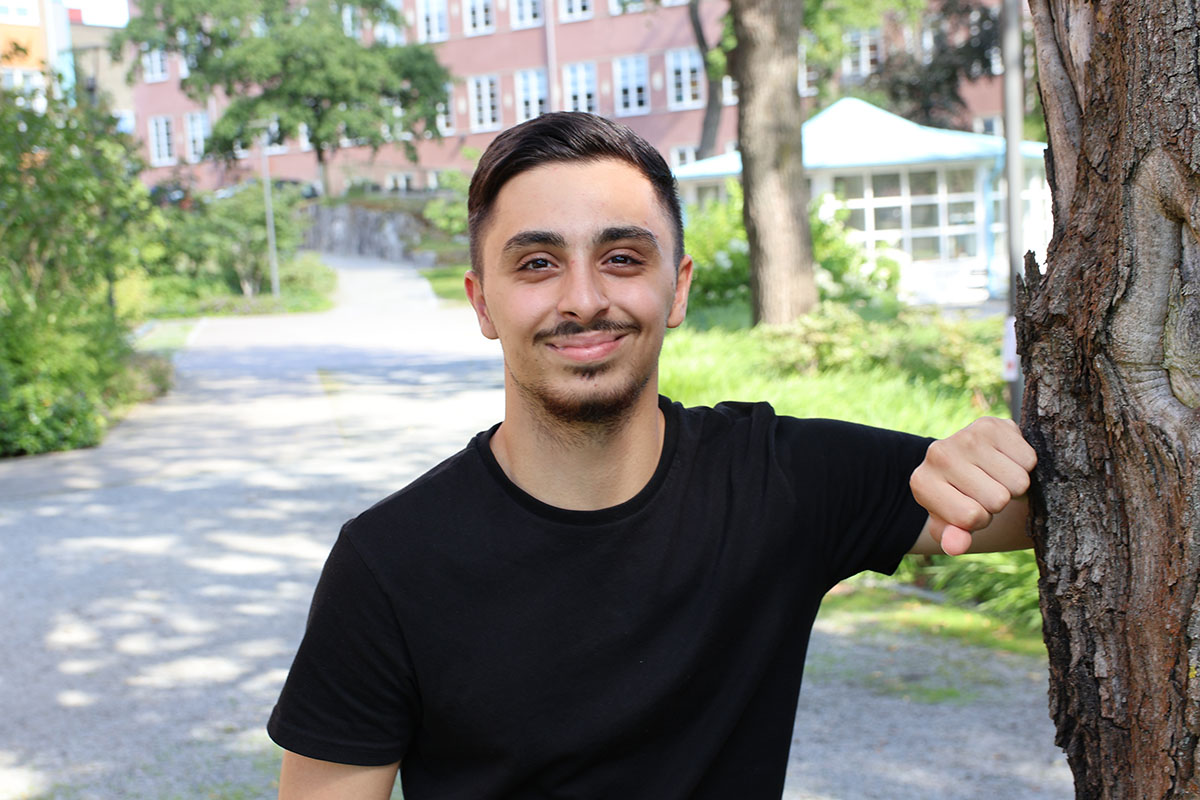 Ung man i svart t-shirt lutar sig mot ett träd och ler, bakgrund med grönska och byggnader.