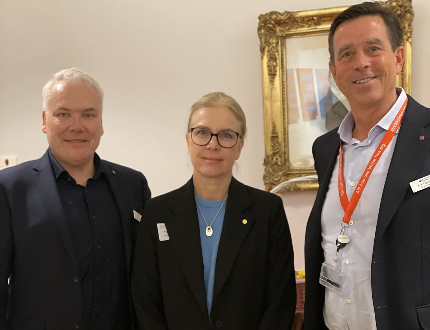 Tre personer i kostym poserar och ler mot kameran, en tavla syns i bakgrunden.