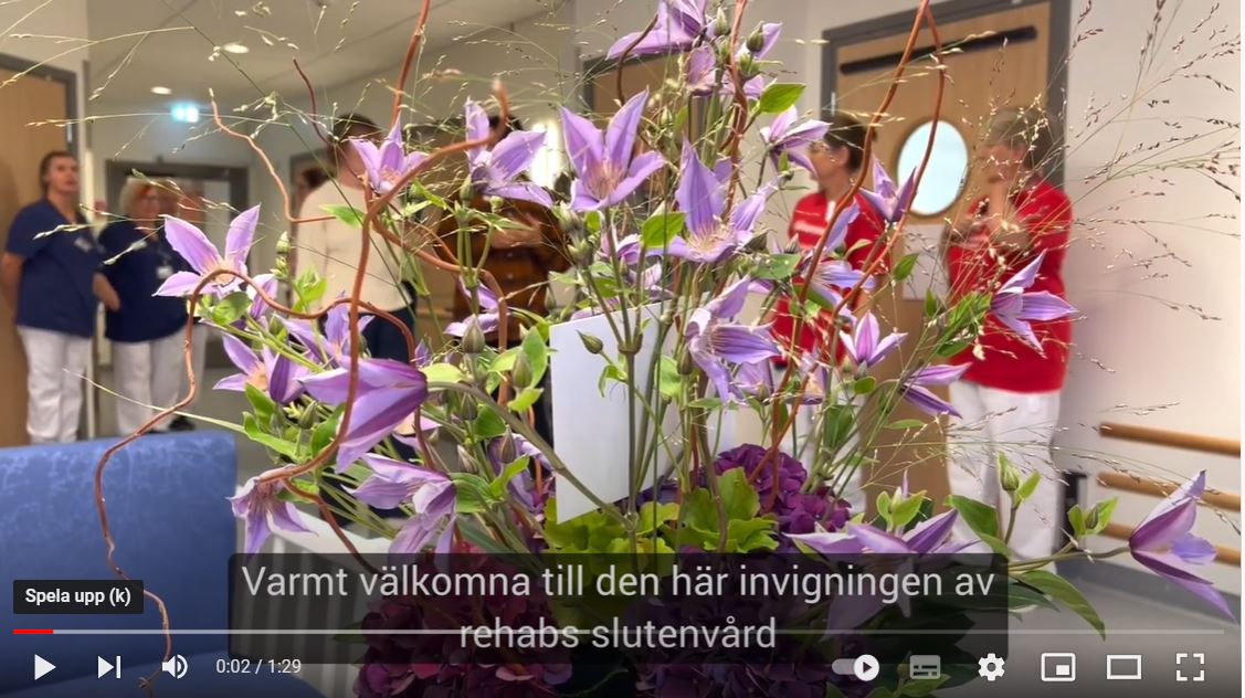 Lila blommor i fokus, personal i färgglada kläder i bakgrunden, text om invigning av rehabs slutenvård.