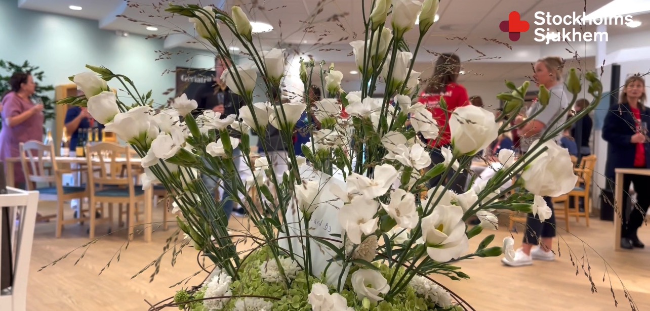 Vit blomsterbukett i fokus med människor i bakgrunden i ett rum med Stockholms Sjukhem-logga upptill.