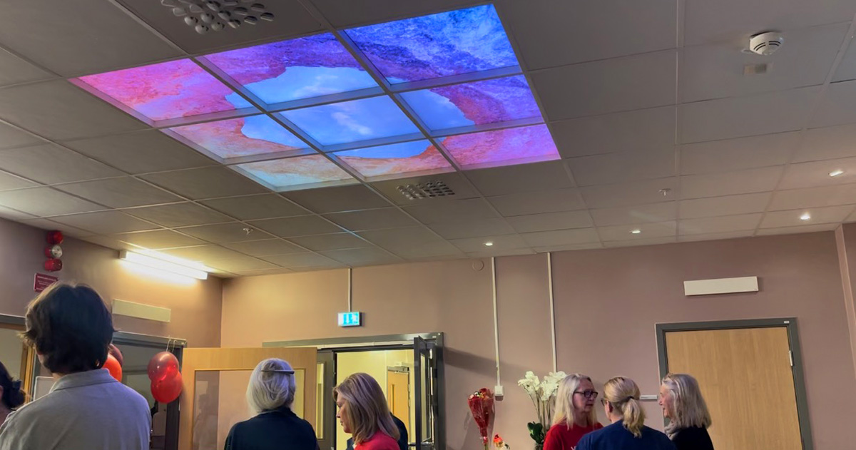 Flera personer pratar i ett rum, med ett takfönster som visar en himmel med moln och färgade kanter.