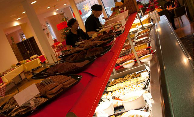 Julbord med olika maträtter uppdukade på buffébord, två personer bakom bordet, julgran i bakgrunden.
