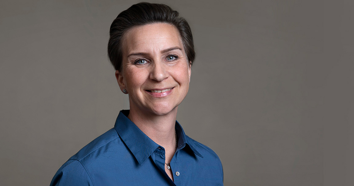 Leende kvinna med kort hår och blå skjorta mot grå bakgrund.