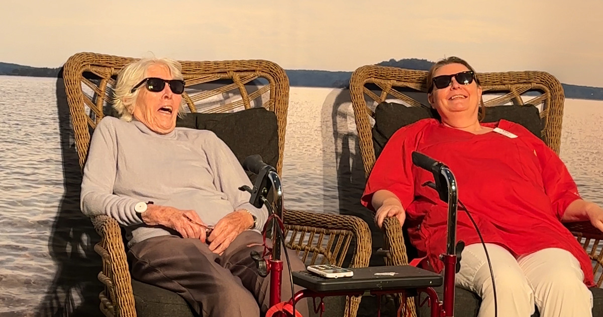Två kvinnor med solglasögon sitter och skrattar i korgstolar vid en sjö i solnedgång.