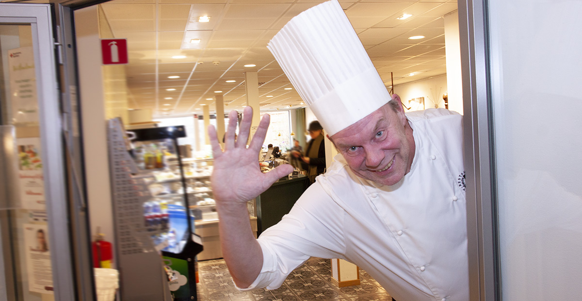 Kock i vit uniform och kockmössa vinkar och ler bredvid en dörr på en restaurang eller café.