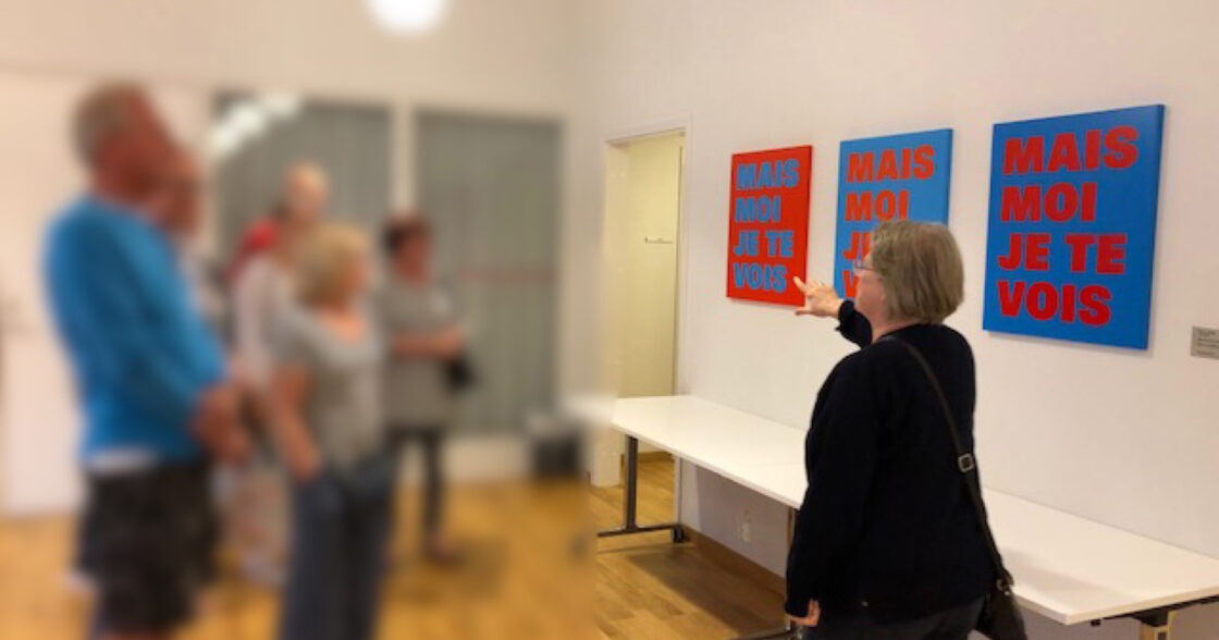 Folk betraktar färgglada tavlor med texten "MAIS MOI JE TE VOIS" på väggen i ett galleri.